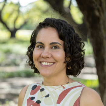 Woman with short curly hair smiling outdoors, wearing patterned sleeveless top near trees.